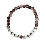 A beaded stretch bracelet on a plain light background. The bracelet features faceted dark brown and black beads with clear inclusions, alternating with white round letter beads spelling 'DEEP ROOTS'.