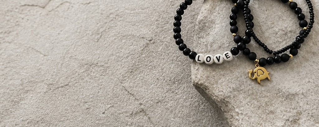 Two beaded stretch bracelets on a light stone surface. One bracelet has white letter beads spelling MAYTH and LOVE, and the other has black beads with a small gold elephant charm.