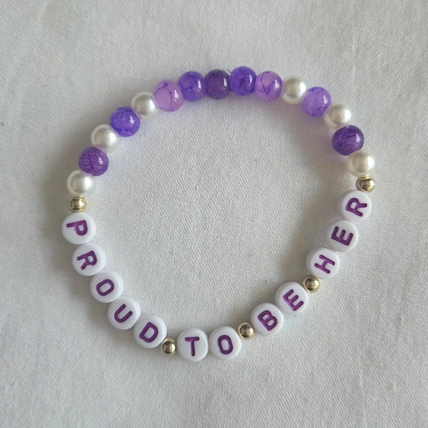 Proud to Be Her bracelet with purple, white, and gold beads on soft fabric background