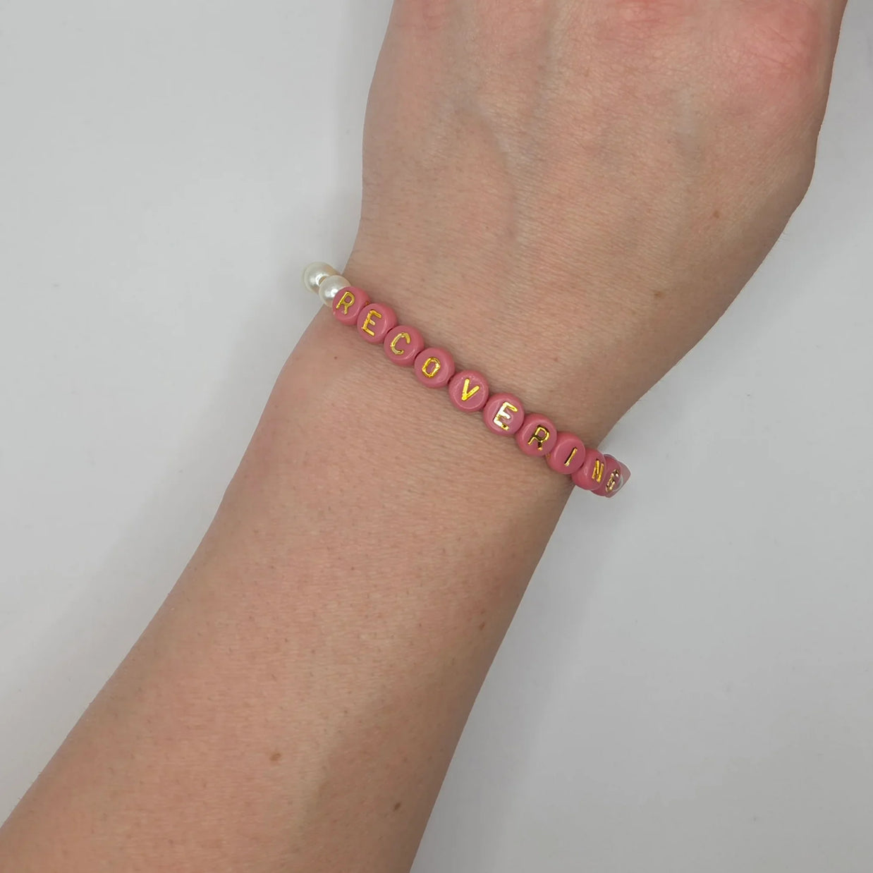 Pink beaded bracelet spelling 'RECOVERING' with gold letters and pearl accents, worn on wrist