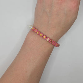 Pink beaded bracelet spelling 'RECOVERING' with gold letters and pearl accents, worn on wrist