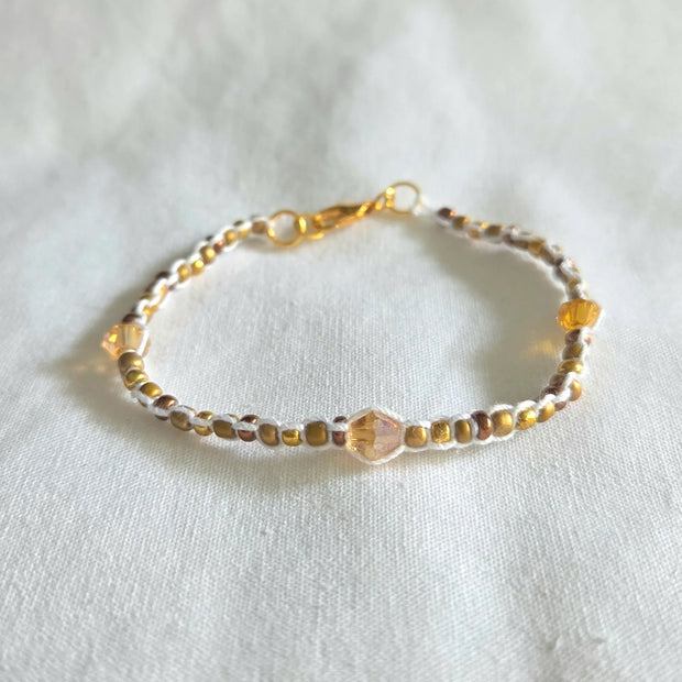Handmade beaded bracelet with gold and amber crystals, white cord, and gold clasp on white fabric