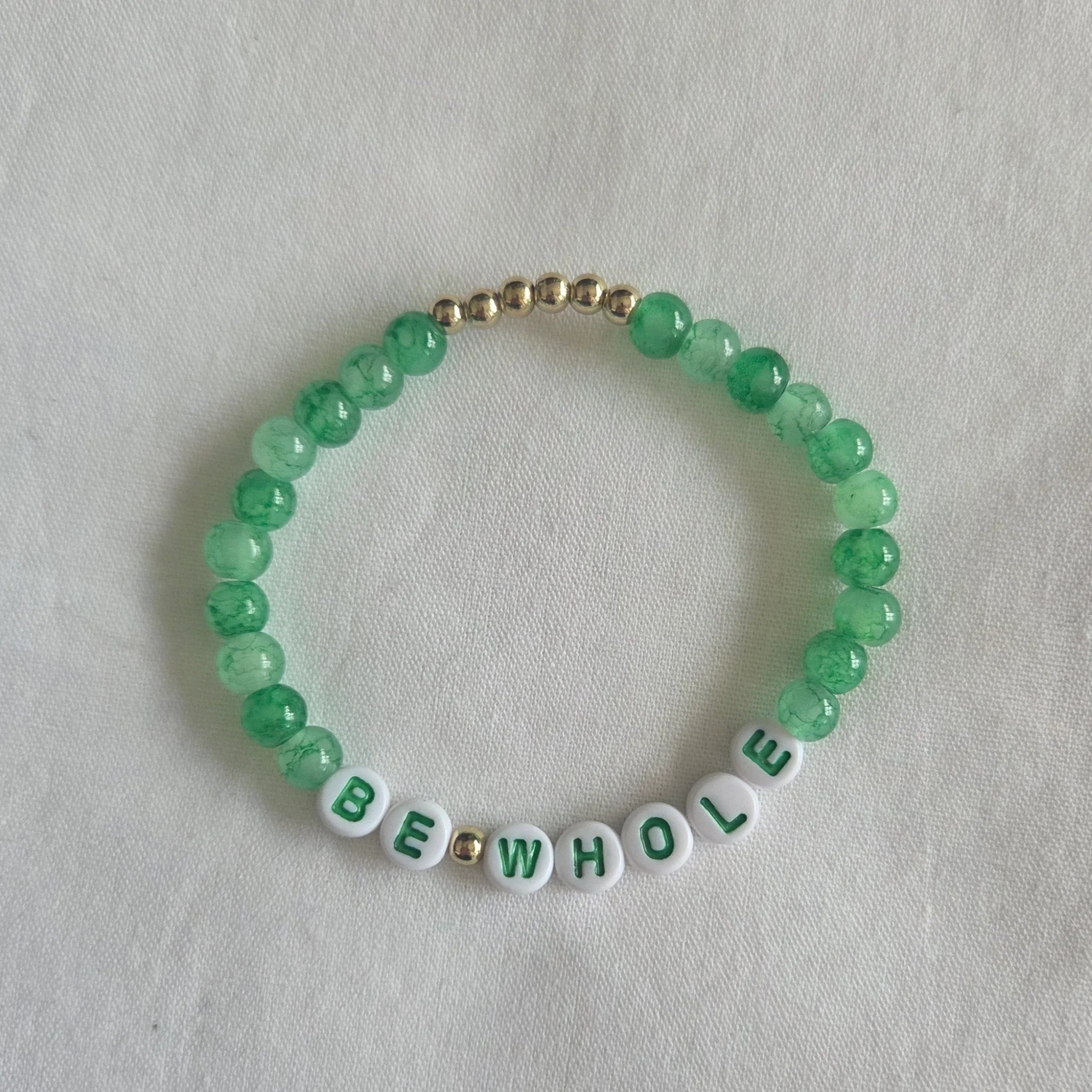 Green beaded bracelet with 'BE WHOLE' letter beads and gold accents on white fabric
