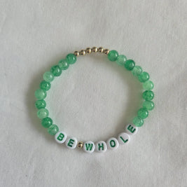Green beaded bracelet with 'BE WHOLE' letter beads and gold accents on white fabric