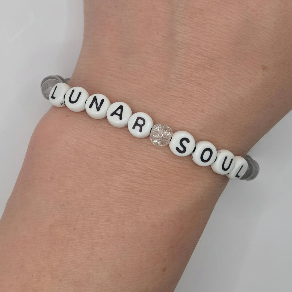 A beaded bracelet worn on a wrist, featuring white round beads with black lettering that spell 'LUNAR SOUL' and a small clear faceted crystal charm in the center, set against a light gray cord.