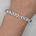 A beaded bracelet worn on a wrist, featuring white round beads with black lettering that spell 'LUNAR SOUL' and a small clear faceted crystal charm in the center, set against a light gray cord.