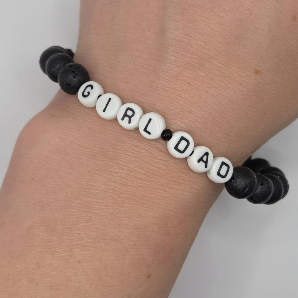 A beaded bracelet worn on a wrist, featuring white round letter beads spelling 'GIRL DAD' with black lettering, separated by small black beads, and connected to black lava stone beads on both sides.