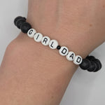 A beaded bracelet worn on a wrist, featuring white round letter beads spelling 'GIRL DAD' with black lettering, separated by small black beads, and connected to black lava stone beads on both sides.