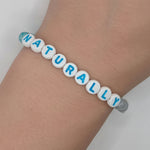 A beaded bracelet spelling 'NATURALLY' in blue letters on white round beads, worn on a person's wrist against a light gray background.