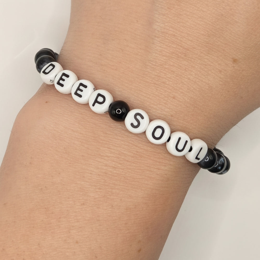 A beaded stretch bracelet worn on a wrist, featuring white letter beads spelling 'DEEP SOUL' with black lettering, accented by black marbled beads on both ends.
