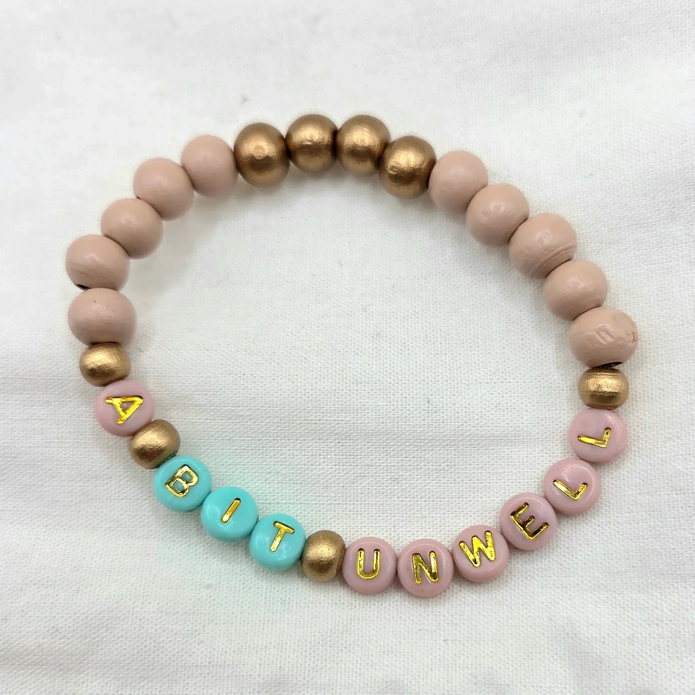 Handmade beaded bracelet with 'A BIT UNWELL' in gold letters on pink and teal beads, perfect for cozy gifts.