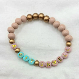 Handmade beaded bracelet with 'A BIT UNWELL' in gold letters on pink and teal beads, perfect for cozy gifts.