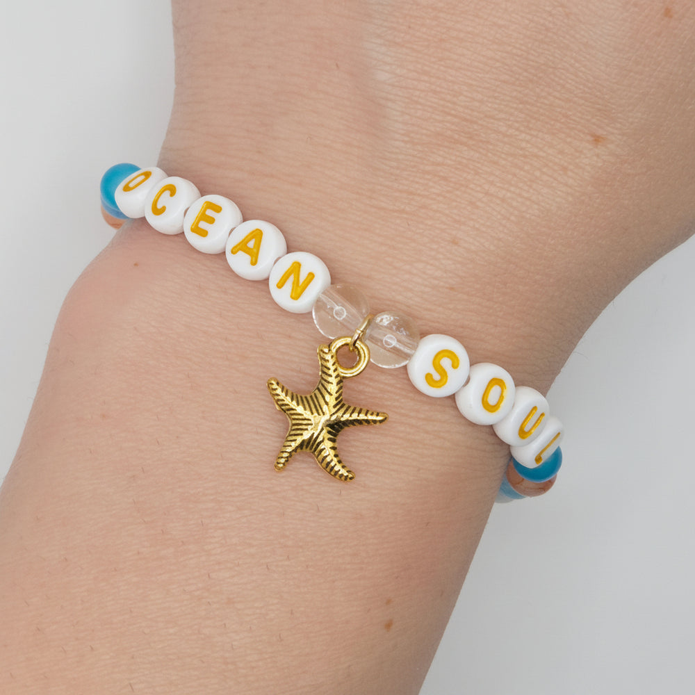 A close-up of a person's wrist wearing a beaded bracelet with white letter beads spelling 'OCEAN SOUL', blue accent beads, clear spacer beads, and a gold starfish charm.