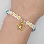 A close-up of a person's wrist wearing a beaded bracelet with white letter beads spelling 'OCEAN SOUL', blue accent beads, clear spacer beads, and a gold starfish charm.