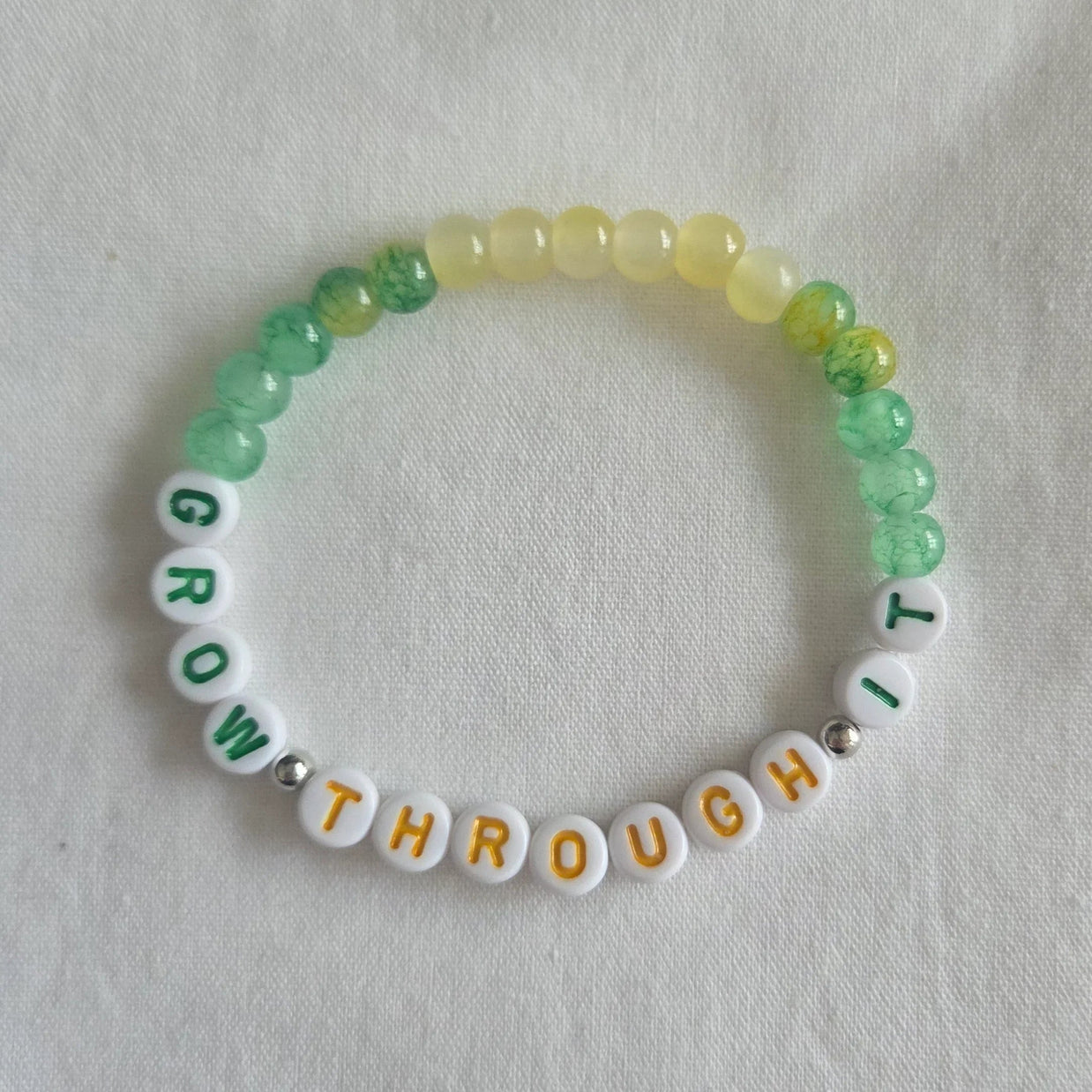 Handmade 'Grow Through It' beaded bracelet with green, yellow, and white beads on a soft fabric background