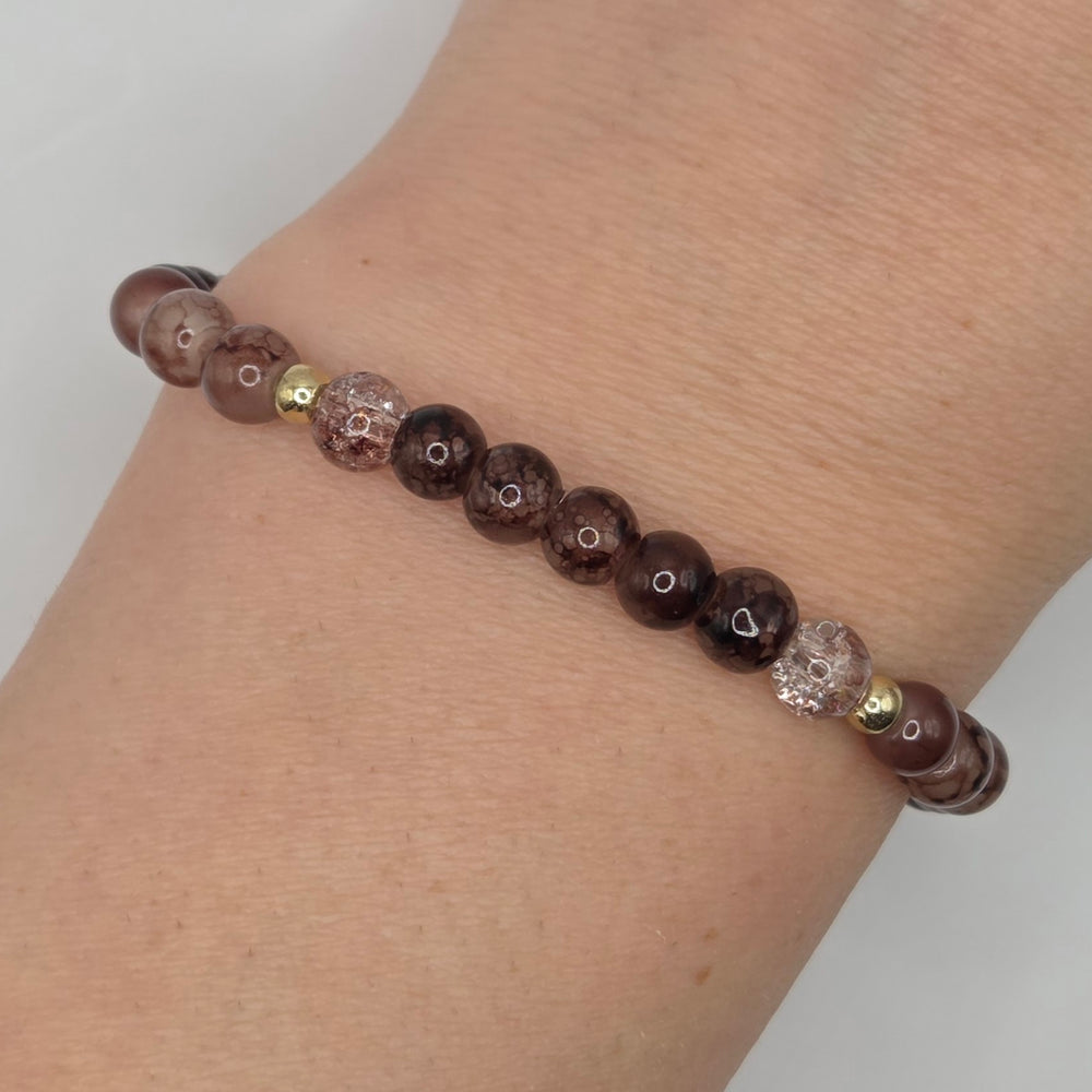A beaded stretch bracelet worn on a wrist, featuring dark brown and black marbled round beads, clear faceted beads with heart details, and small gold spacer beads.