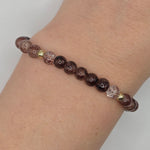 A beaded stretch bracelet worn on a wrist, featuring dark brown and black marbled round beads, clear faceted beads with heart details, and small gold spacer beads.