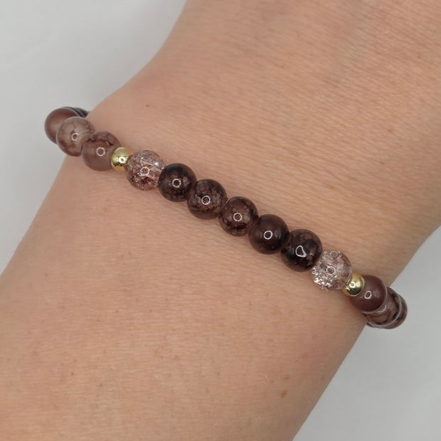 A beaded stretch bracelet worn on a wrist, featuring dark brown and black marbled round beads, clear faceted beads with heart details, and small gold spacer beads.