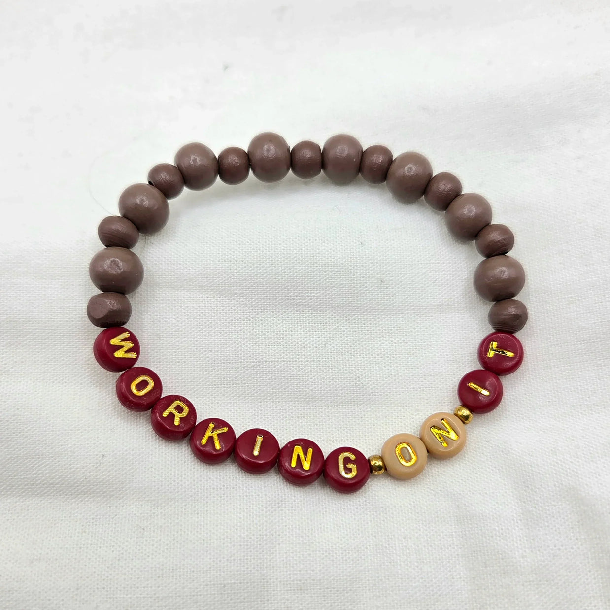 Handmade 'WORKING ON IT' beaded bracelet with maroon, brown, and gold letter beads on white fabric
