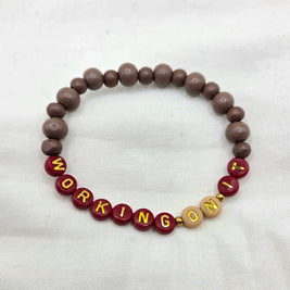Handmade 'WORKING ON IT' beaded bracelet with maroon, brown, and gold letter beads on white fabric
